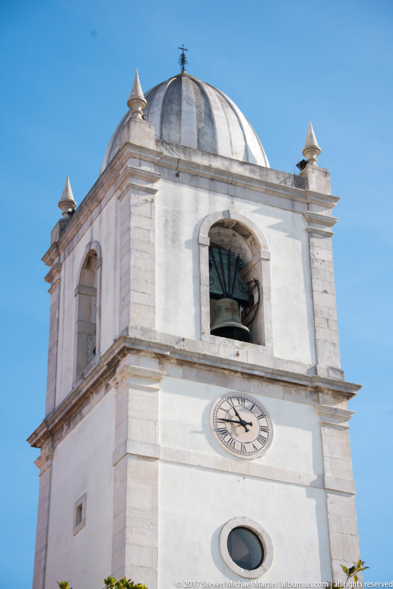 Close view of a white bell tower with arched bell opening, round clock, circular window, domed roof and corner finials.