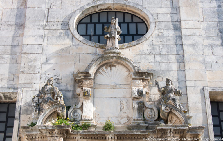 Weathered stone wall detail with oval window, central shell niche, scrolling ornament and several carved religious figures.