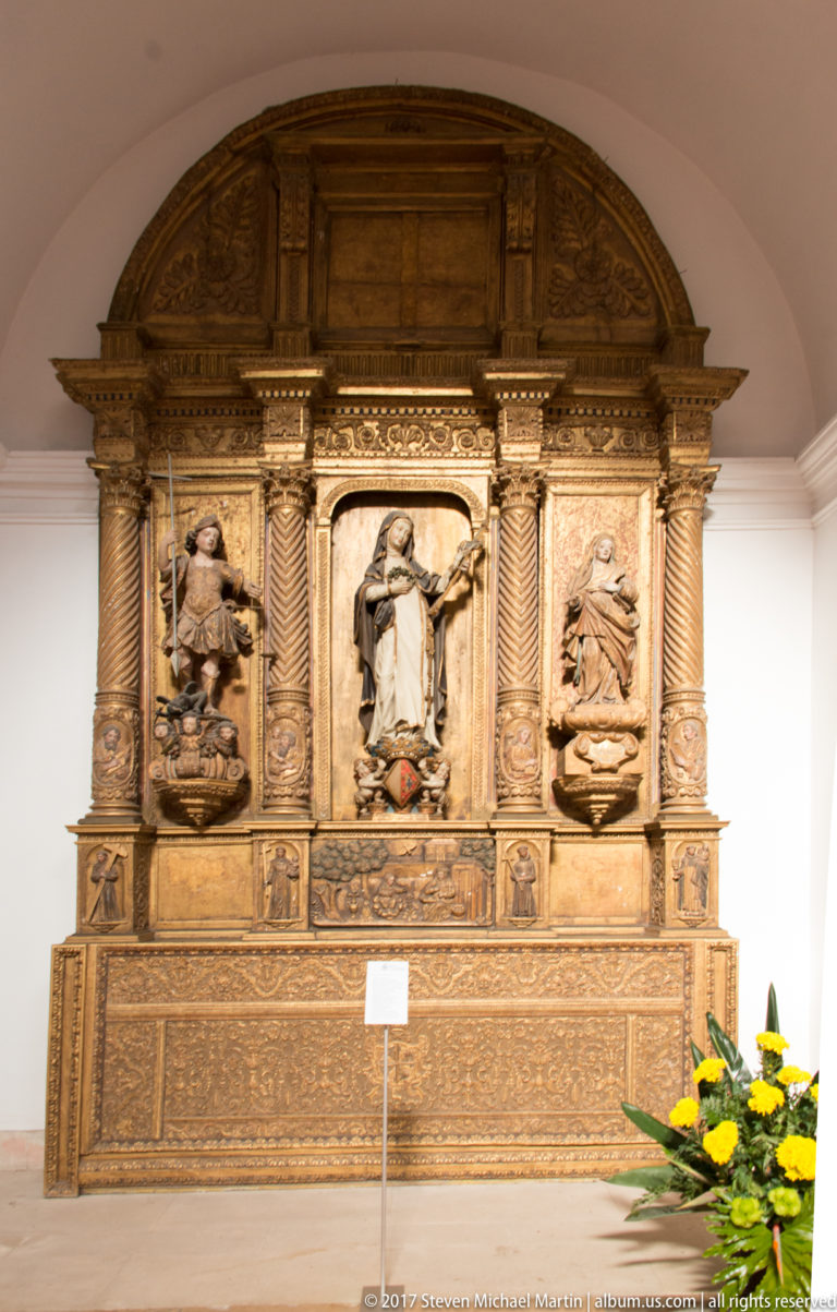 Large dark carved wooden retable with three religious sculptural panels, twisted columns and a broad arched top.