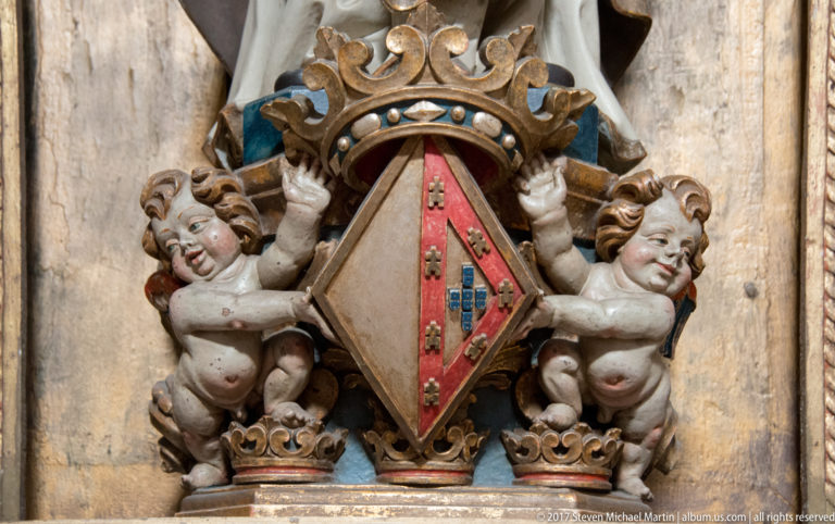 Close view of two carved putti supporting a crowned red-and-tan heraldic shield with traces of blue and gold paint.