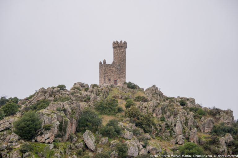 Crenellated stone tower standing alone on a rocky, scrub-covered hill beneath a gray sky.