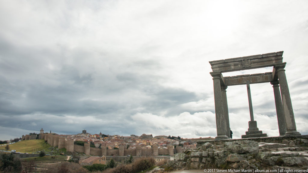 Four-column stone cross monument on rocky ground overlooking the distant walled city of Ávila.
