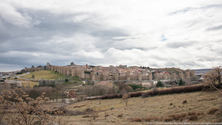 Wide view of Ávila’s walls and towers across brown open fields beneath a cloudy sky.