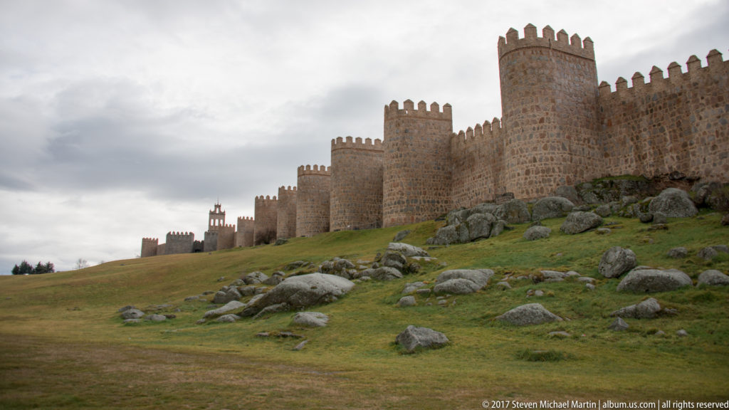 Low-angle view of Ávila’s crenellated towers above a grassy slope scattered with large rocks.