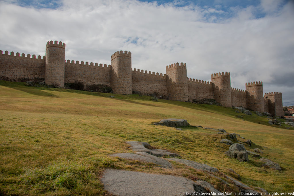 Long crenellated wall with repeated stone towers extending across a broad grassy foreground in Ávila.