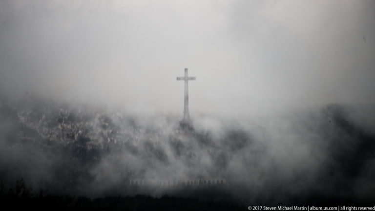 Tall cross silhouetted in dense fog above a dark hillside with barely visible structures below.