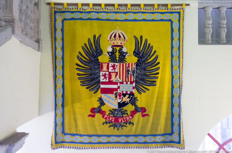 Yellow heraldic banner with black double-headed eagle, crowned shield and “PLVS VLTRA” motto.