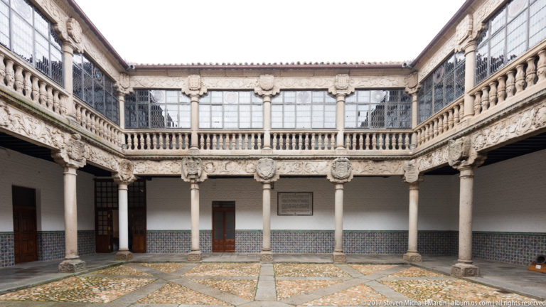 Ornate courtyard with stone columns, carved shields, glazed upper gallery, balustrades and patterned paving.