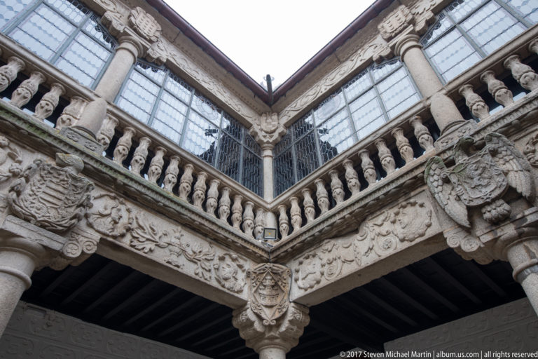 Low-angle detail of carved stone heraldry, corbel, balustrade and glazed upper windows in a courtyard.