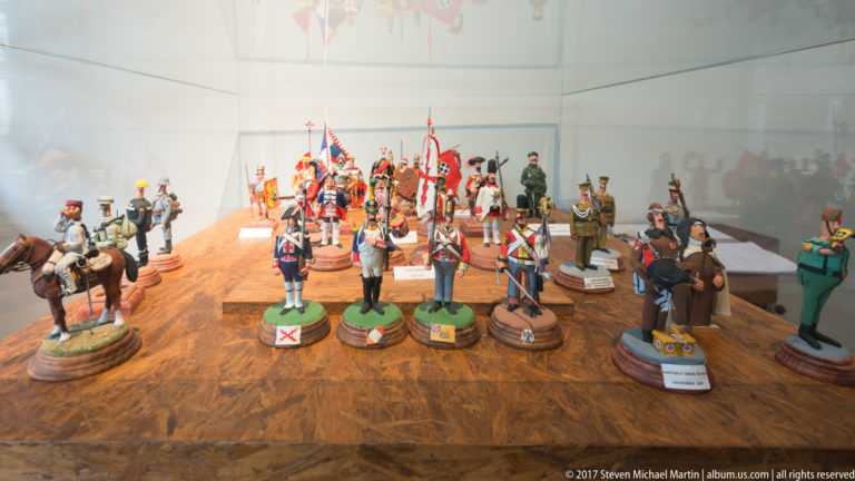 Painted miniature soldiers, cavalry and flag bearers arranged in rows inside a glass museum case.