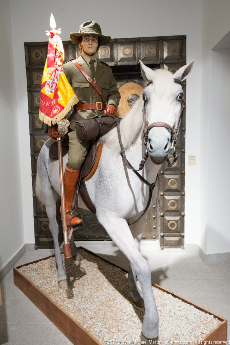 Uniformed military mannequin riding a white horse mount and carrying a red-and-yellow heraldic standard in a museum display.