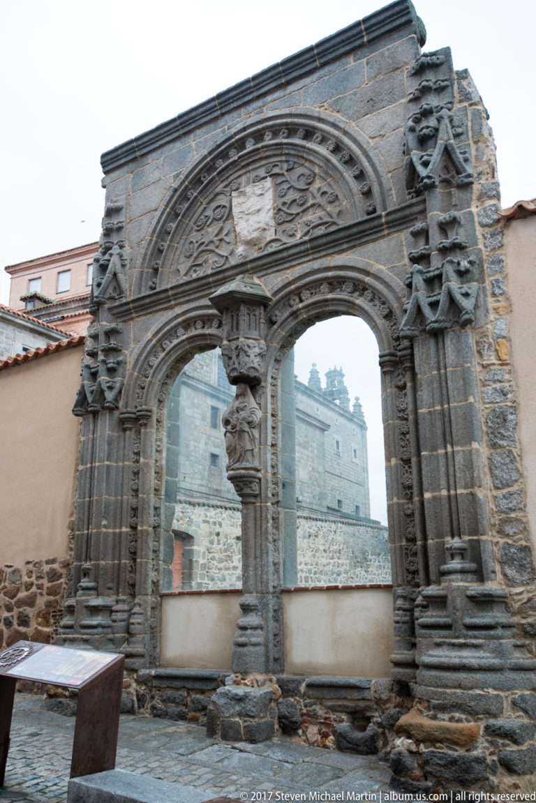 Ornately carved dark-stone double portal with relief decoration standing before masonry buildings.