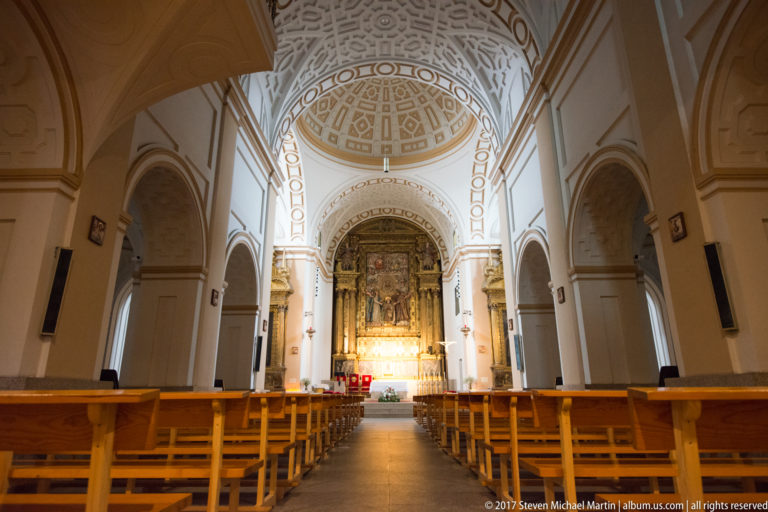 Nave of the Basilica of Saint Teresa in Ávila, with wooden pews leading toward the illuminated high altar.