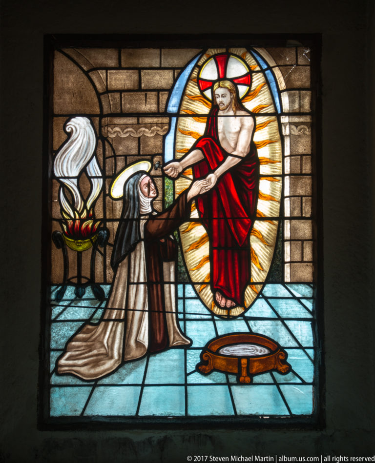 Stained glass showing Saint Teresa of Ávila kneeling and reaching toward a radiant figure of Christ.