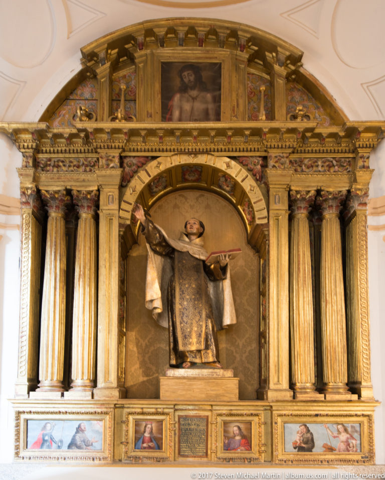 Robed friar statue holding an open book in a gilded niche between tall fluted columns.