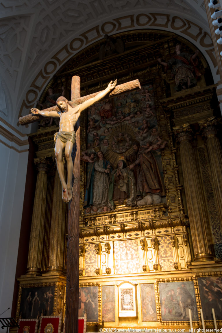 Large wooden crucifix with carved Christ figure standing before an ornate gilded church retable.