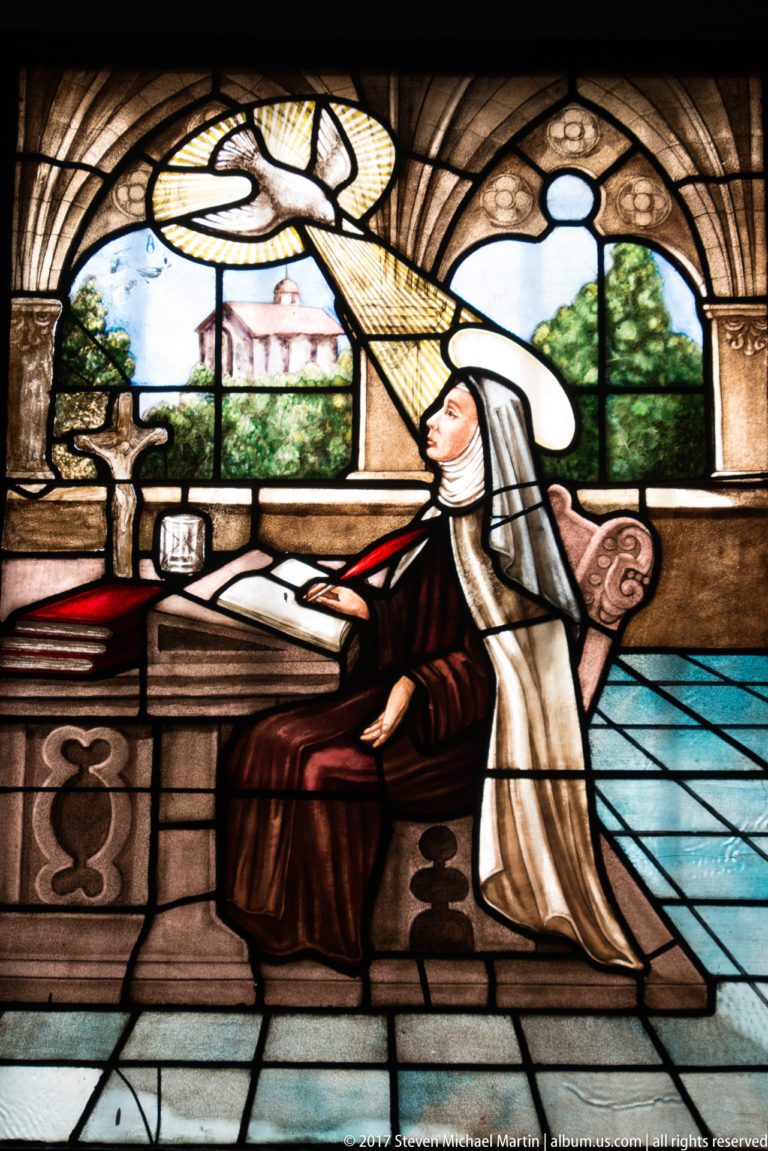 Saint Teresa of Ávila in Carmelite habit seated at a desk with a manuscript beside an open window.