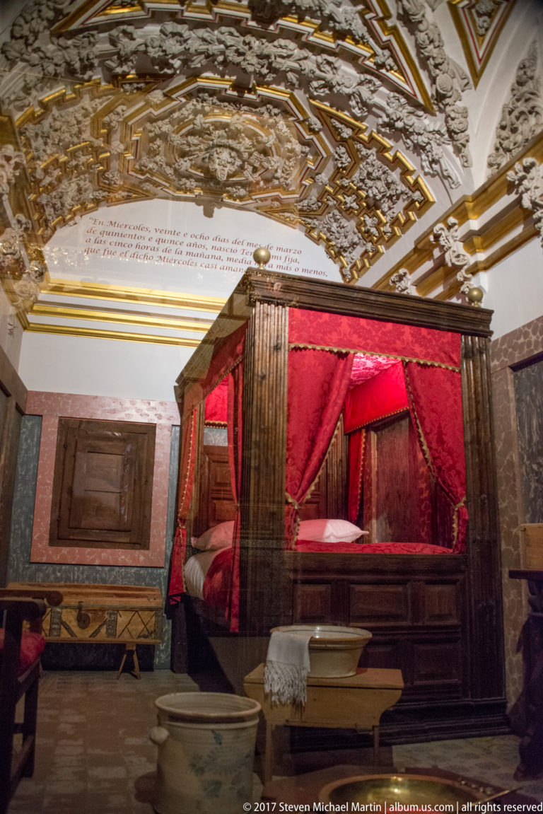 Canopied wooden bed with red curtains in a historic-room display at the Saint Teresa birthplace complex in Ávila.