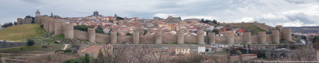 Panoramic view of Ávila’s crenellated walls enclosing dense red-roofed buildings on a hill.