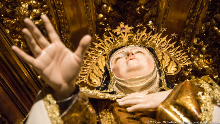 Close low-angle view of Saint Teresa of Ávila raising one hand against a richly gilded background.
