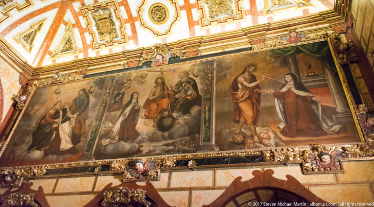 Two large religious paintings from a Saint Teresa narrative cycle mounted high beneath an ornate ceiling.