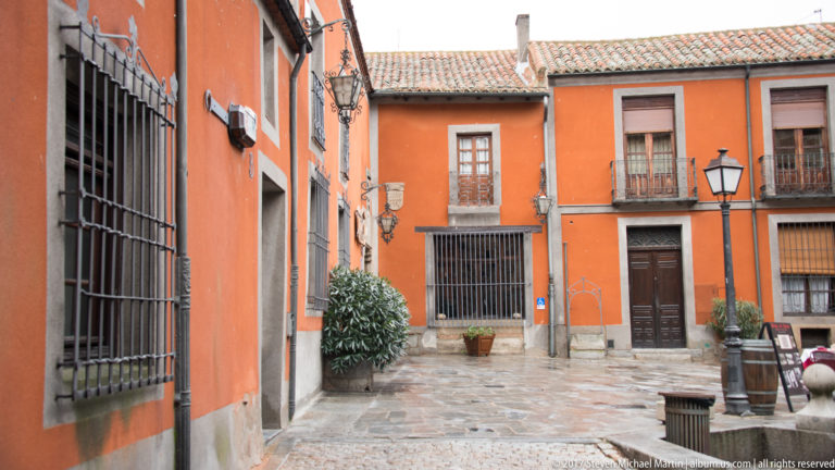 Small stone-paved courtyard enclosed by orange façades with barred windows, balconies, doors and potted plants.