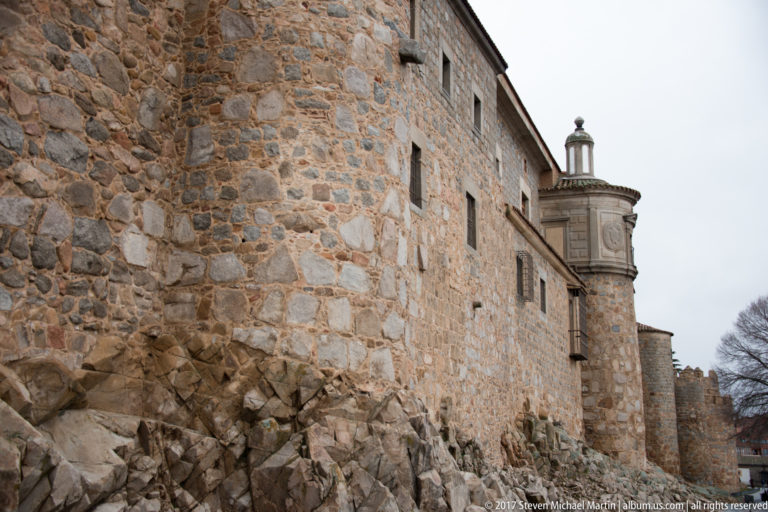 Long rough-stone defensive wall rising from exposed bedrock with small openings and a projecting rounded structure.