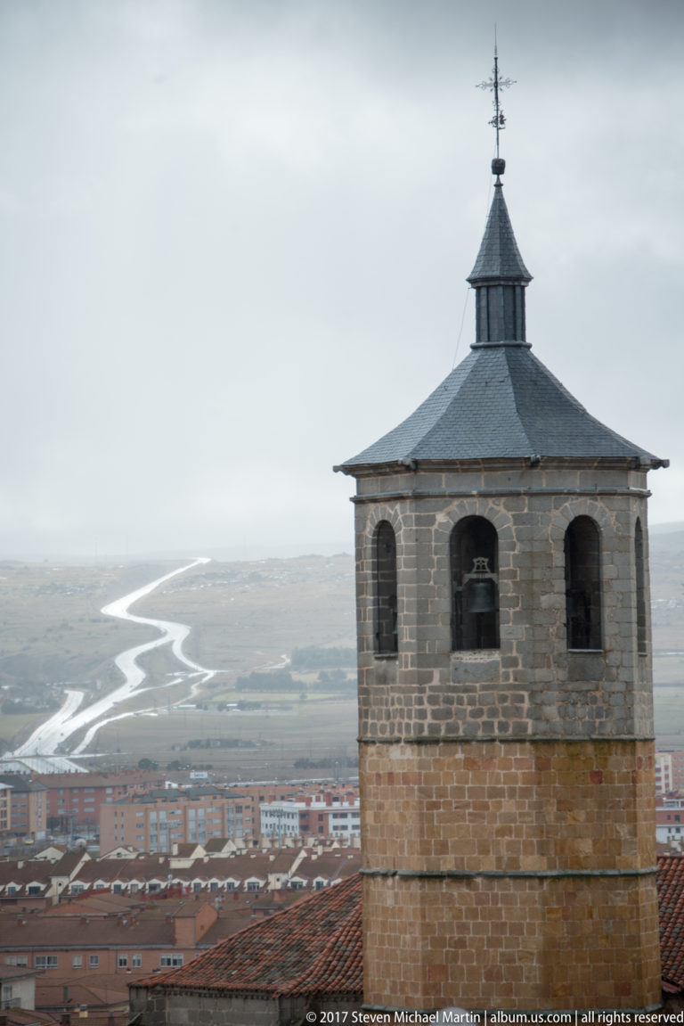Square stone bell tower with slate roof and weather vane above rooftops and a broad misty plain.