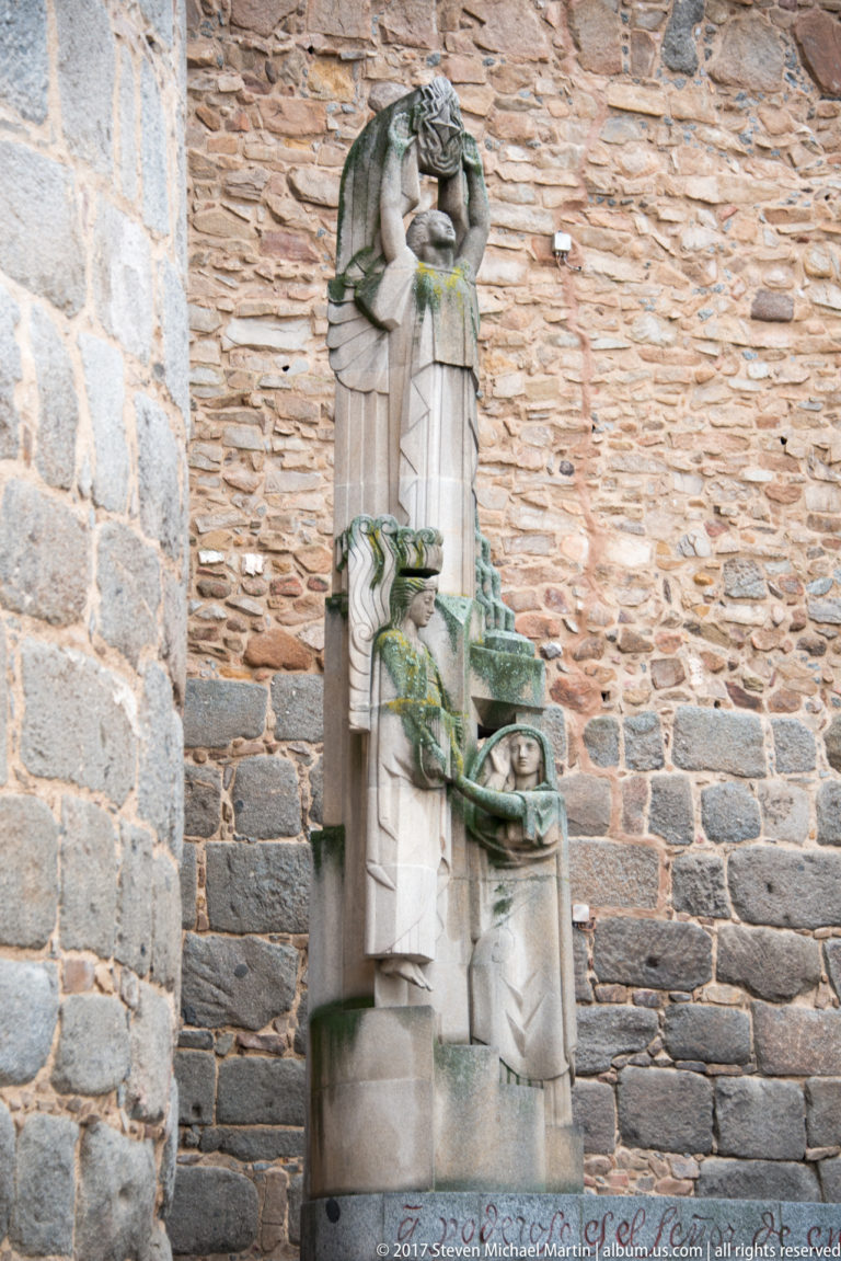 Tall pale-stone monument with stylized carved figures standing against a rough historic masonry wall.