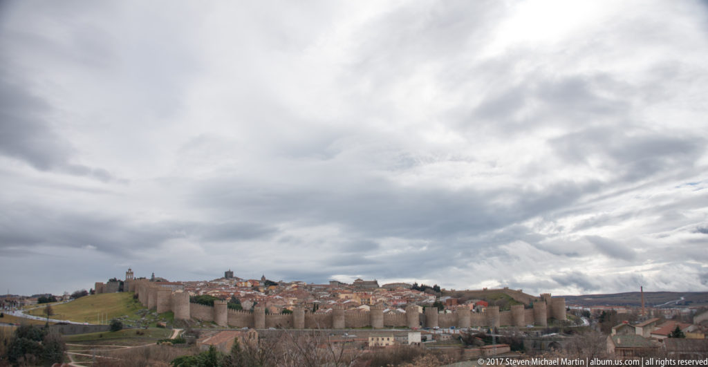 Distant walled city of Ávila beneath dramatic layered storm clouds, with open land in the foreground.
