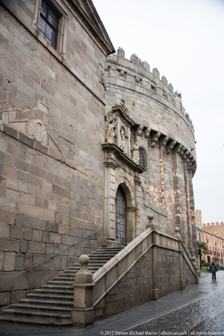 Stone staircase leading to an ornate doorway beneath a crenellated parapet in a massive masonry wall.