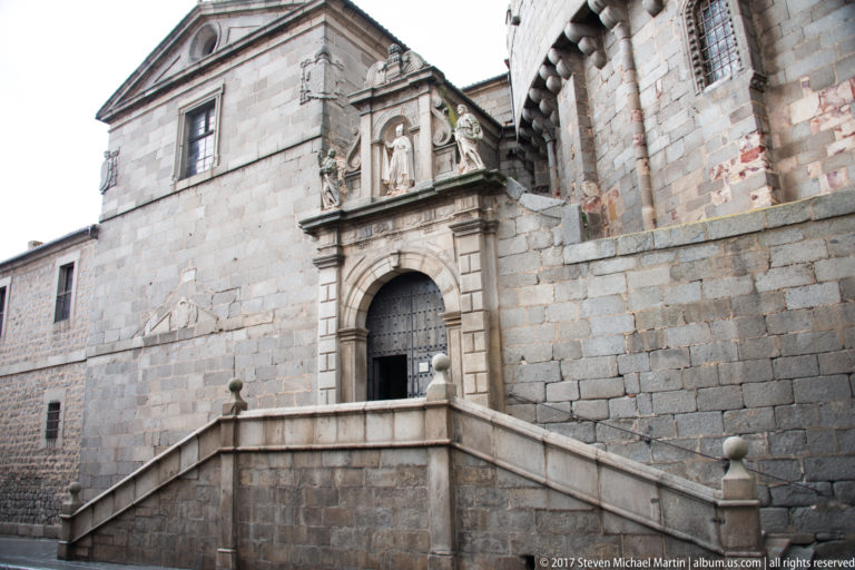 Split stone staircase rising to an ornate portal with a saint statue set in a broad masonry façade.