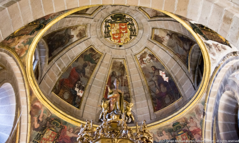Stone church dome divided into painted religious panels with gilded ribs and a heraldic shield near the crown.