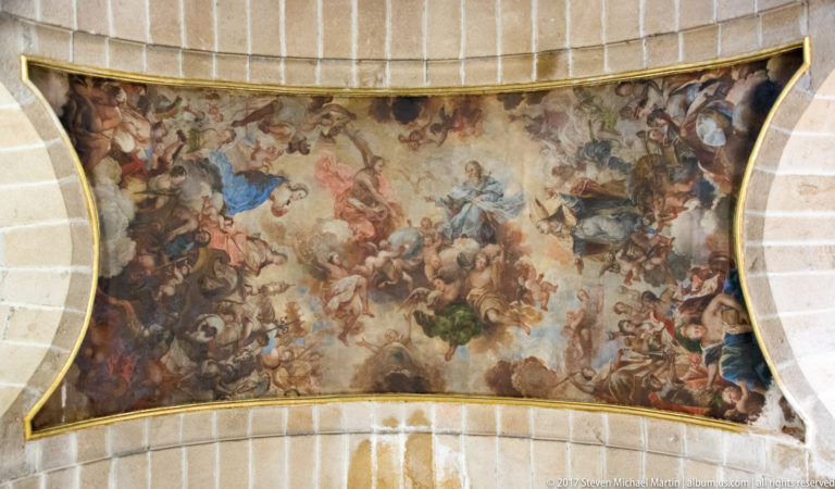 Large crowded religious fresco with many figures and clouds filling a curved ceiling panel between pale stone ribs.