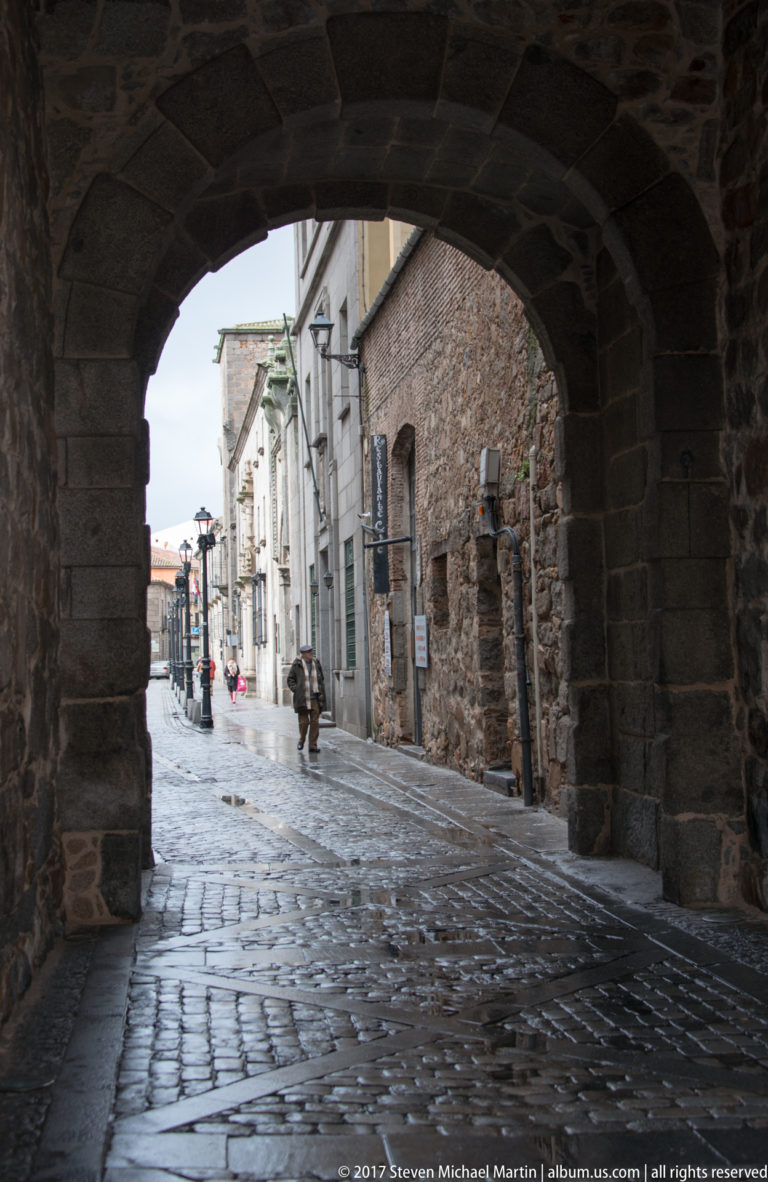 Wet cobbled street and pedestrians seen through the dark opening of a massive stone archway in Ávila.