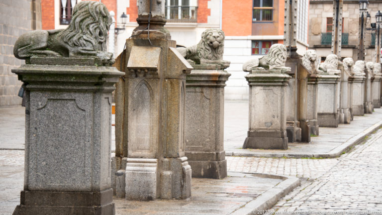 Row of square stone posts, several topped by carved crouching lions, receding along a paved plaza.