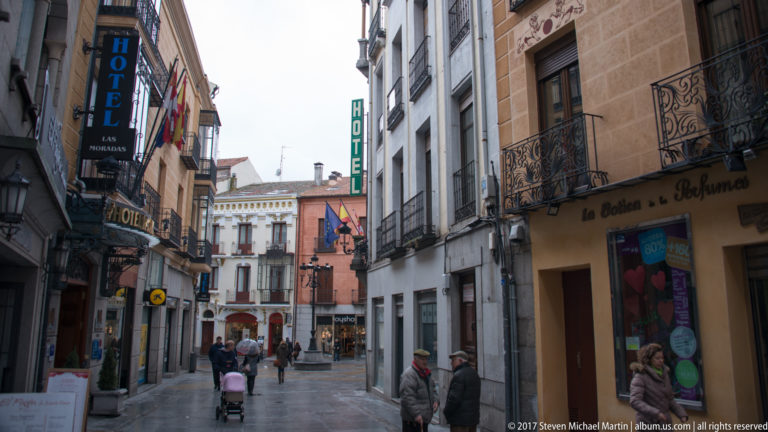 Pedestrians on a narrow historic street lined with hotel signs, balconies, flags and multi-story façades.