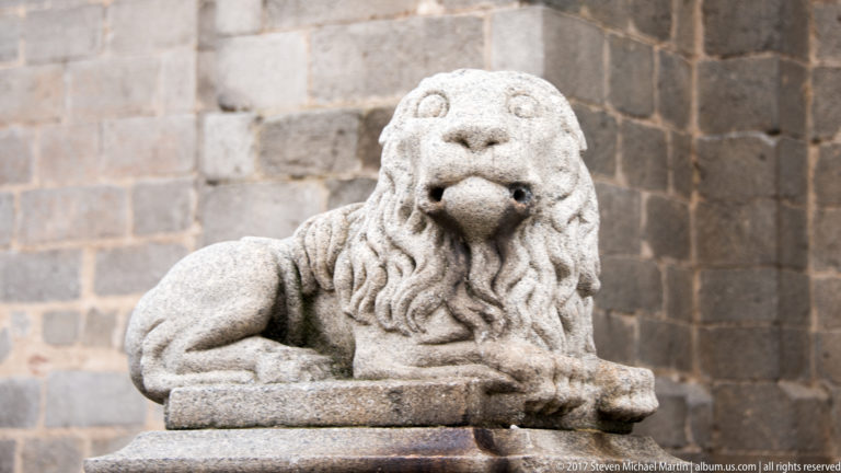 Weathered recumbent stone lion with carved mane and paws on a pedestal before a block-stone wall in Ávila.