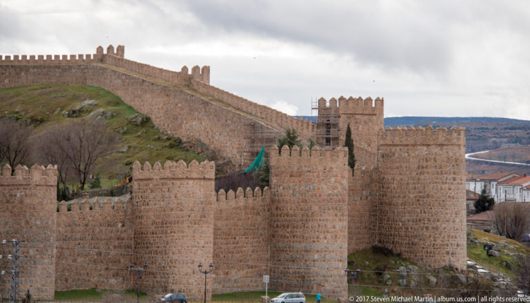 Crenellated stone walls and projecting towers intersect and descend across a grassy hillside in Ávila.