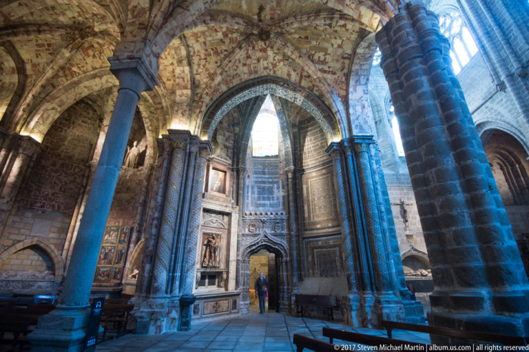Stone side chapel with thick columns, patterned vaulting, wall monuments, painted panels, and cool daylight from tall windows at right.