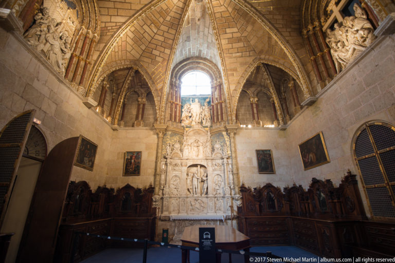 Tall carved stone monument centered beneath a round window, with dark wooden stalls and framed paintings along the chapel walls.