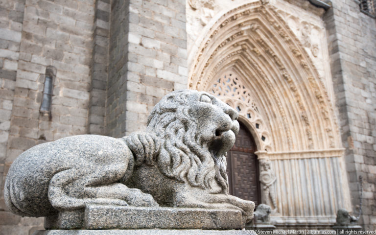 Recumbent stone lion in the foreground before a deeply recessed pointed Gothic portal with multiple carved archivolts.