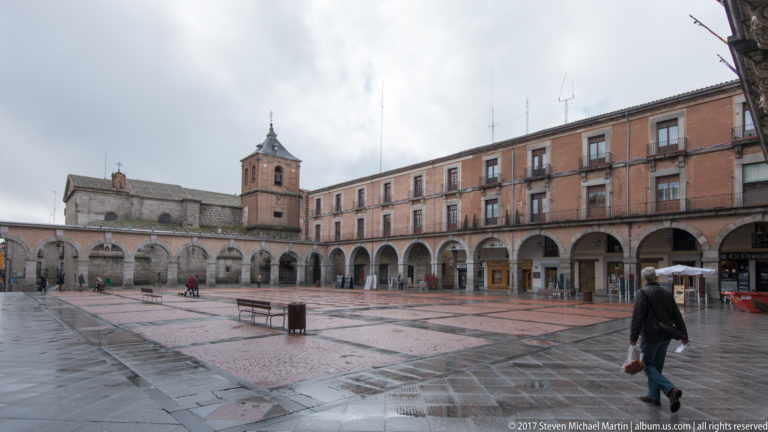 Wet paved civic square lined by long arcaded buildings and balconies, with a church tower at the far side and scattered pedestrians.