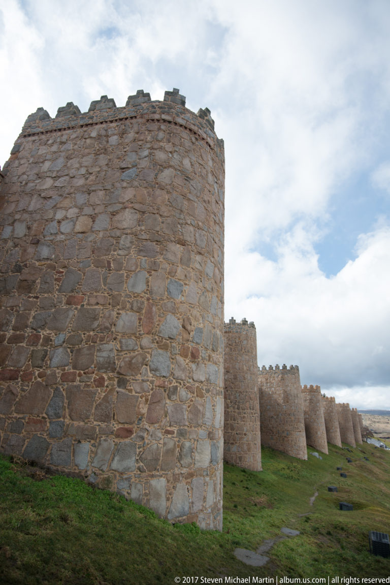 Row of round stone towers and crenellated curtain wall receding across a grassy slope beneath a cloudy sky in Ávila.