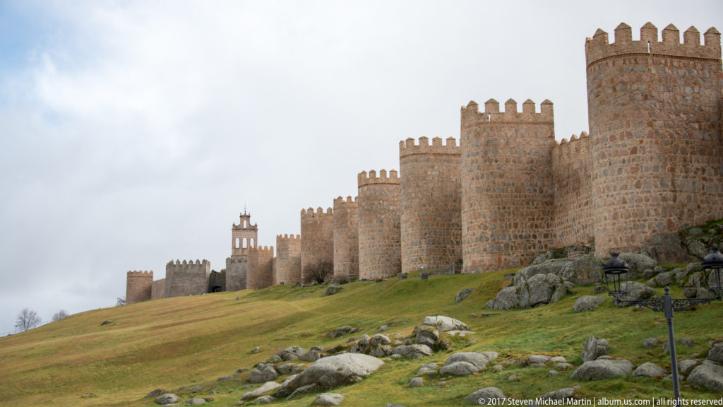 Long crenellated stone wall with repeated projecting towers climbing across a grassy hillside, seen from near a large foreground tower.