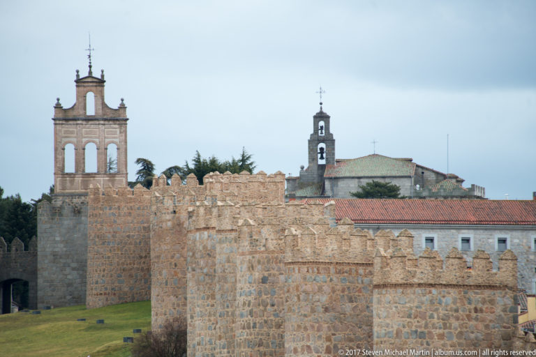 Stone battlements and a large wall tower in front of two church bell towers in Ávila.