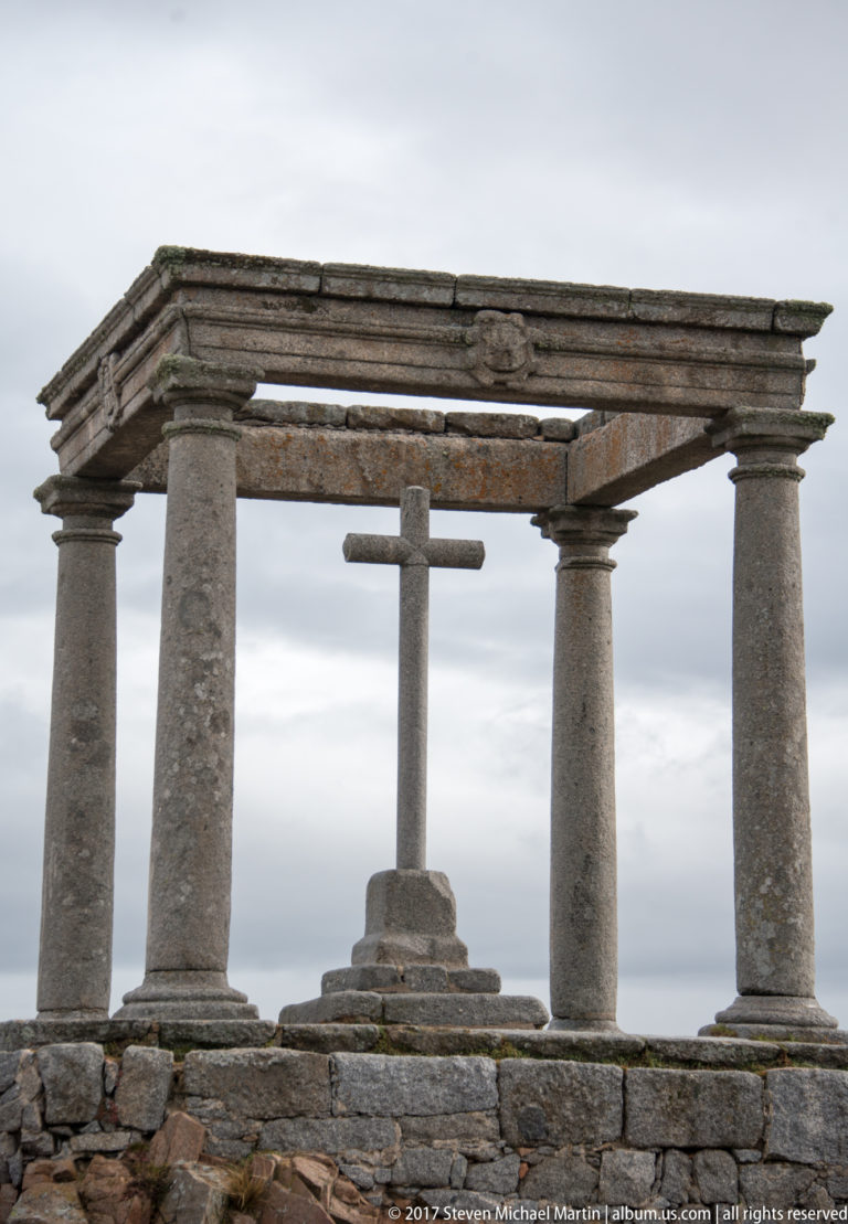 Stone cross on a stepped pedestal beneath a rectangular four-column stone canopy against a cloudy sky.