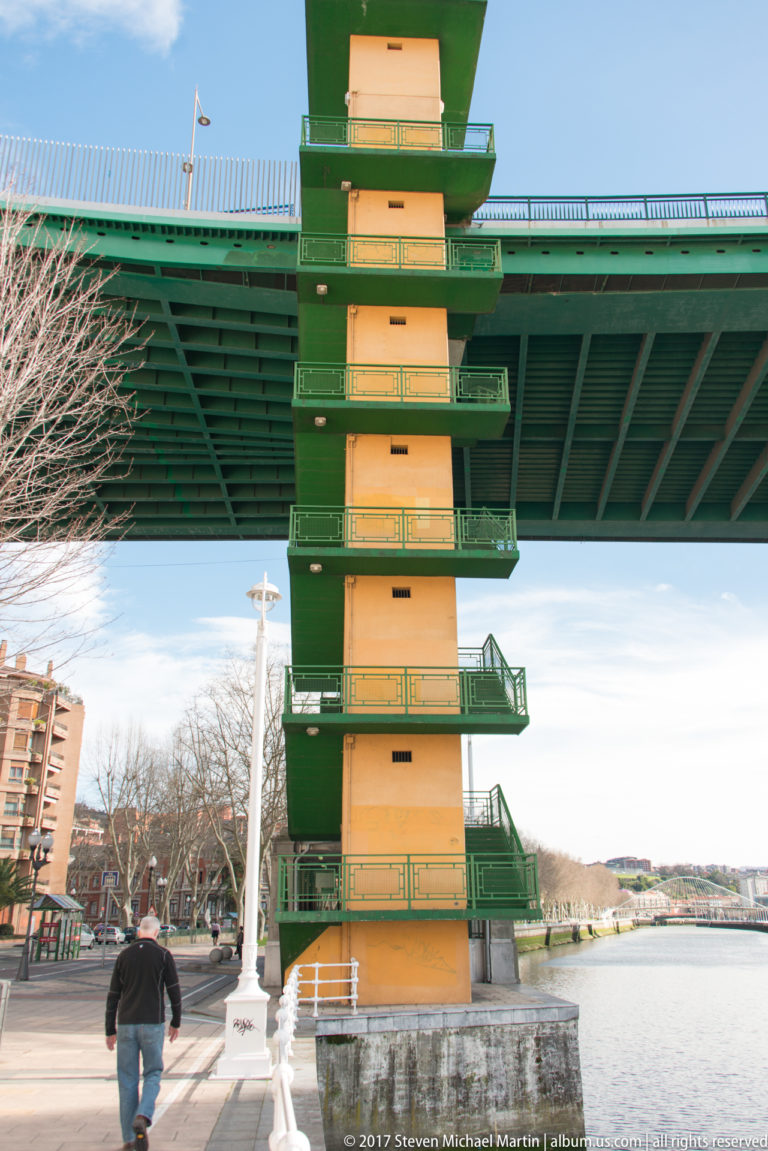 Yellow stair tower with green landings descending beneath La Salve Bridge beside the riverside promenade.
