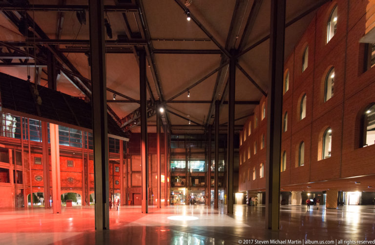 Wide Azkuna Zentroa atrium with black steel columns, roof trusses, red-lit brick walls, arched windows, and reflective flooring.