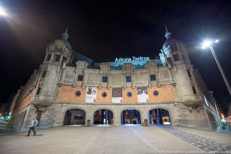 Frontal night view of Azkuna Zentroa with three large arches, ornate corner towers, banners, and its illuminated roofline sign.